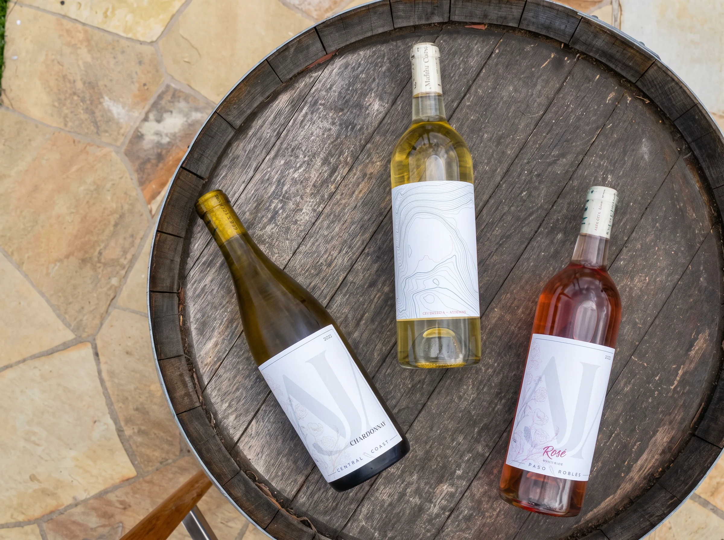 AJA Chardonnay, white blend, and Rose bottles resting on a wine barrel